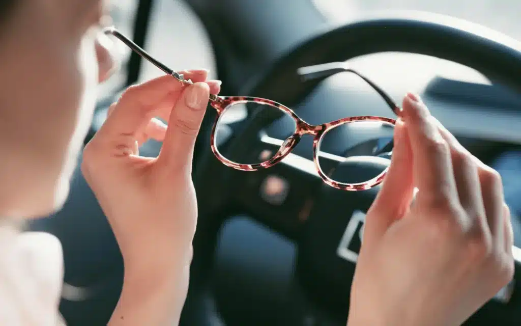 A female holds spectacles in front of a car's steering wheel.
