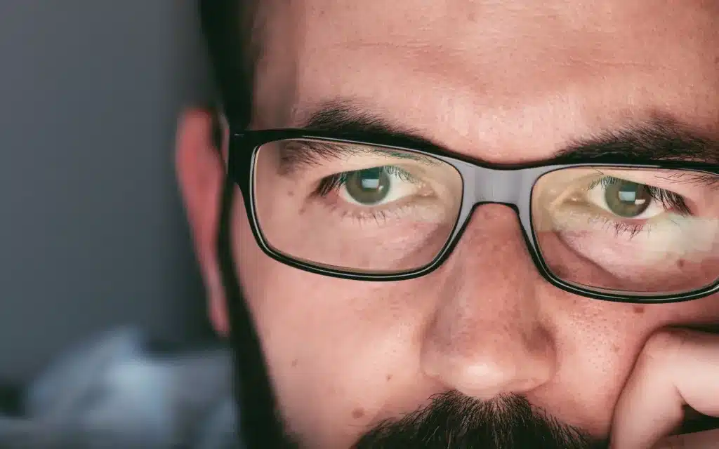 Close up of young adult man in glasses working on laptop computer at night.