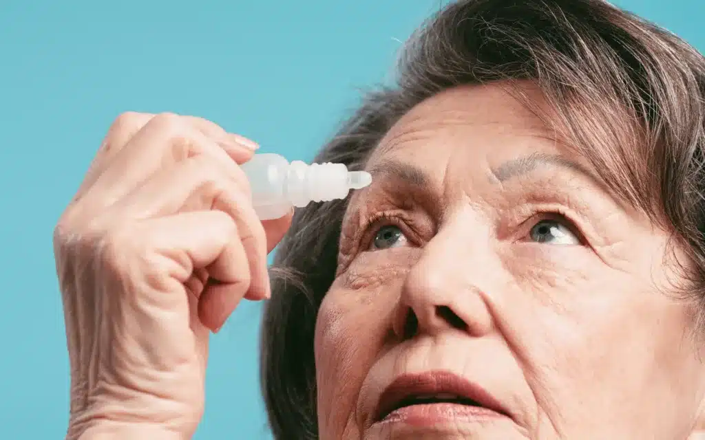 Elderly woman applying eye drops looking up on blue background.
