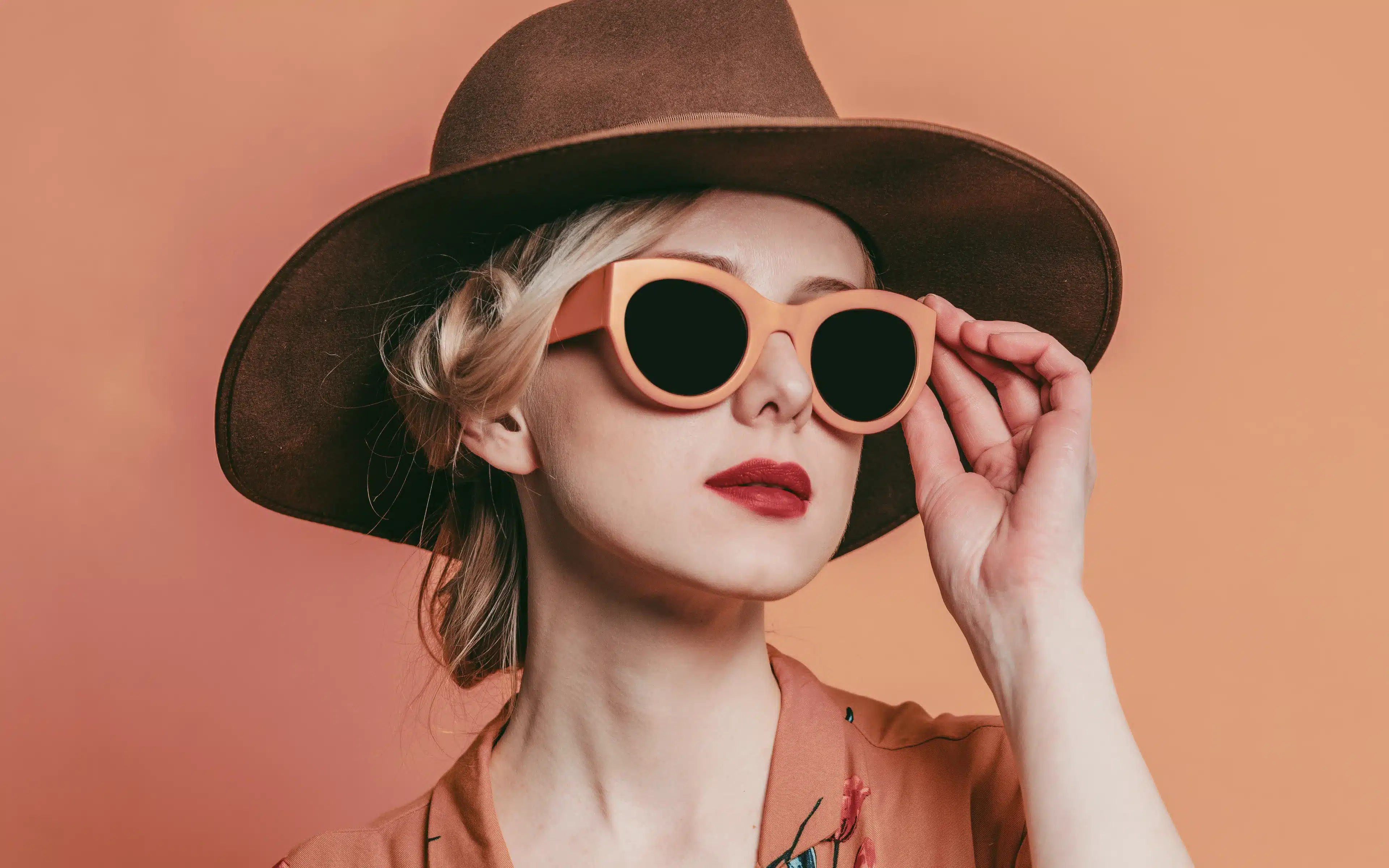 A blonde woman in a wide-brim brown felt hat adjusts oversized round peach sunglasses with dark lenses against a warm terracotta background