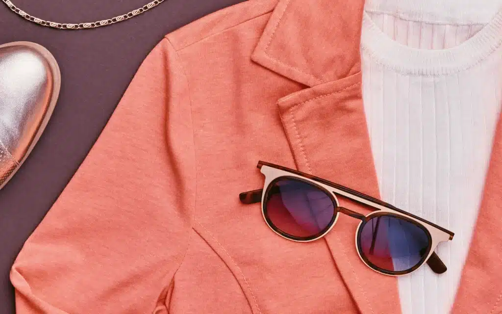 Round tortoiseshell and gold-frame sunglasses with blue-tinted lenses resting on a coral blazer layered over a white ribbed knit top, alongside a silver loafer and gold chain necklace on a purple background