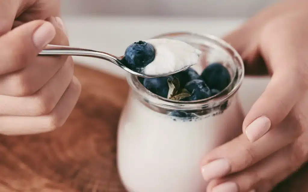 A glass jar of probiotic yoghurt topped with fresh blueberries, with a hand holding a spoon